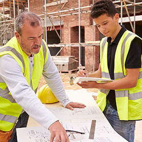 Builder On Building Site Discussing Work With Apprentice